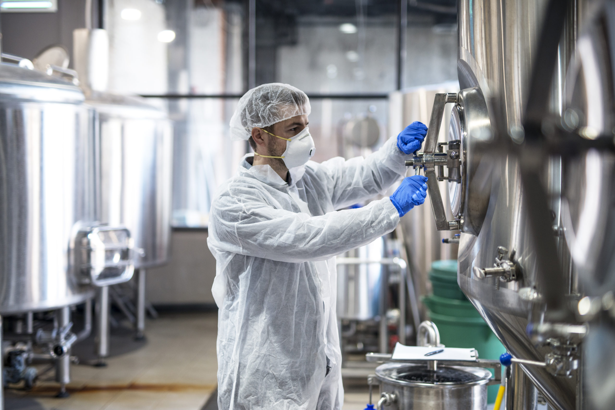 Technologist in protective uniform wearing hairnet mask goggles and gloves closing industrial tank reservoir.